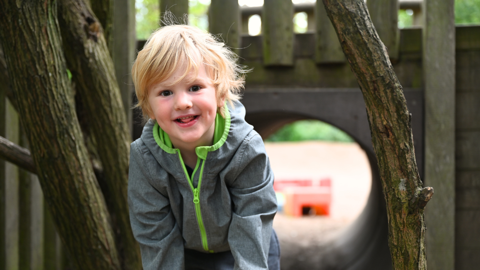 Ein lachender kleiner, blonder Junge sitzt auf einem Baumstamm im Garten vor einem Sandkasten.